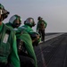 The image shows U.S. Navy flight deck crew members wearing green jerseys, helmets, and protective gear on an aircraft carrier flight deck. Multiple personnel are visible in a line formation, with at least one aircraft visible in the background on the right side. The scene appears to be during active flight deck operations.