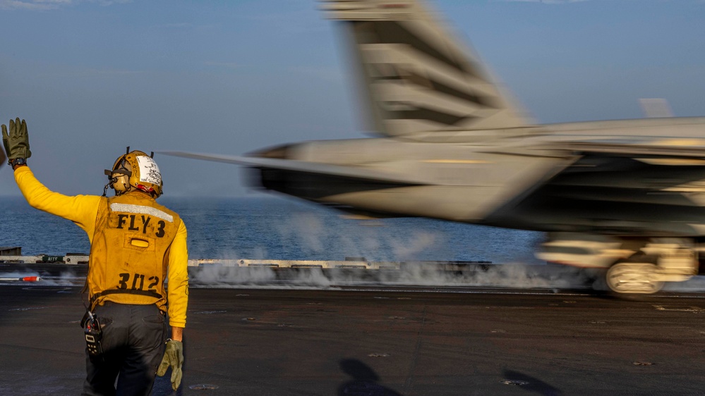A naval flight deck crew member wearing a yellow jersey marked 'FLY 3' and number '302' signals with a raised hand as a military jet aircraft launches from an aircraft carrier catapult. The aircraft is captured in motion blur, indicating high speed during the catapult launch. Steam from the catapult system is visible on the flight deck.