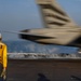 A naval flight deck crew member wearing a yellow jersey marked 'FLY 3' and number '302' signals with a raised hand as a military jet aircraft launches from an aircraft carrier catapult. The aircraft is captured in motion blur, indicating high speed during the catapult launch. Steam from the catapult system is visible on the flight deck.