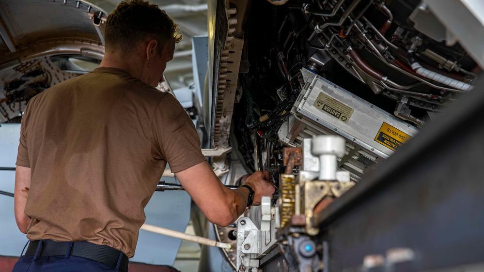 A technician in a tan/brown shirt is performing maintenance work inside an aircraft's internal compartment or weapons bay. The individual is using a hand tool to work on complex mechanical and electrical components. A yellow caution label warning of high voltage (200 volts) is clearly visible on one of the equipment panels.
