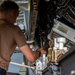 A technician in a tan/brown shirt is performing maintenance work inside an aircraft's internal compartment or weapons bay. The individual is using a hand tool to work on complex mechanical and electrical components. A yellow caution label warning of high voltage (200 volts) is clearly visible on one of the equipment panels.