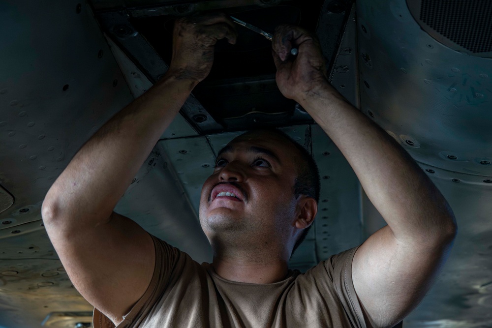 A male technician or mechanic is working underneath a large metal structure, likely an aircraft, using a hand tool. He is looking upward with visible effort on his face, his hands dirty from the work. The image is taken from a low angle, emphasizing the underside of the vehicle and the physical demands of the task.