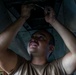 A male technician or mechanic is working underneath a large metal structure, likely an aircraft, using a hand tool. He is looking upward with visible effort on his face, his hands dirty from the work. The image is taken from a low angle, emphasizing the underside of the vehicle and the physical demands of the task.