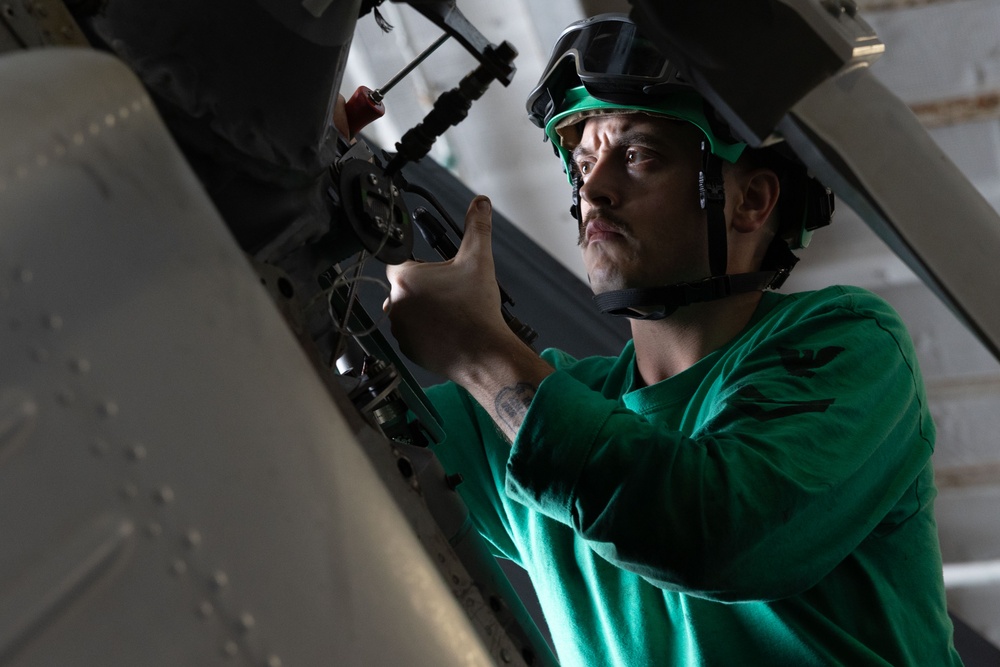 A technician wearing a green jersey and protective helmet with goggles is performing maintenance work on what appears to be a military aircraft. The individual is using a tool to work on mechanical components in a confined access panel area of the aircraft. The green jersey and helmet color are consistent with aviation ordnance or maintenance crew uniforms typically seen on U.S. Navy aircraft carriers.