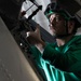 A technician wearing a green jersey and protective helmet with goggles is performing maintenance work on what appears to be a military aircraft. The individual is using a tool to work on mechanical components in a confined access panel area of the aircraft. The green jersey and helmet color are consistent with aviation ordnance or maintenance crew uniforms typically seen on U.S. Navy aircraft carriers.