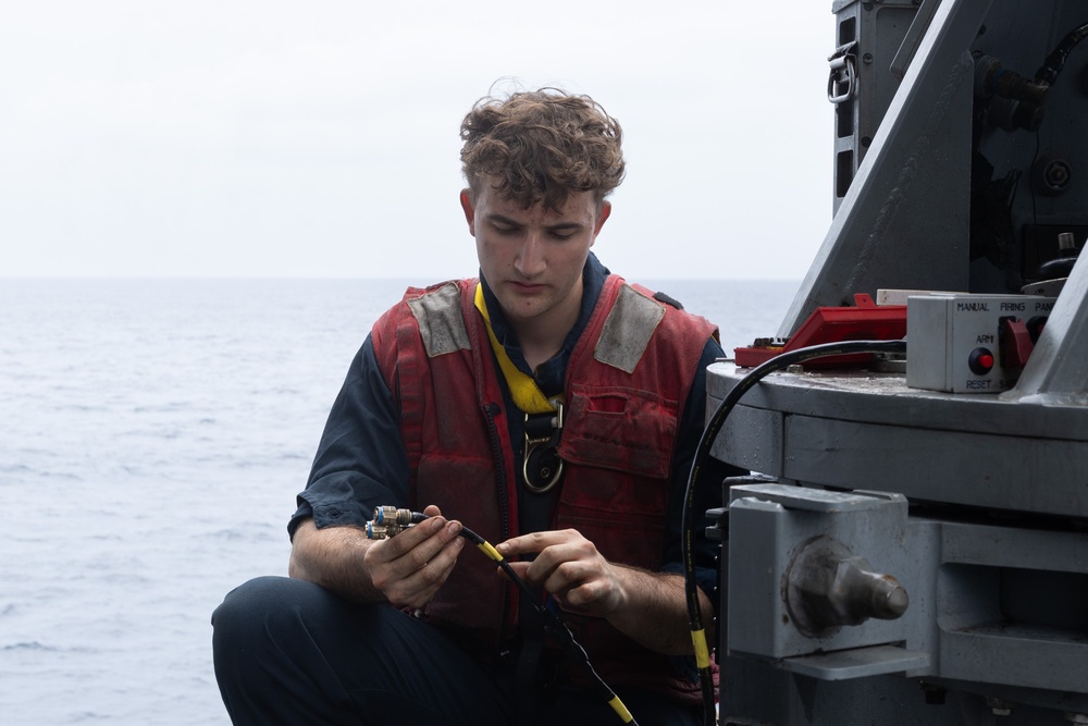 A young male sailor or naval personnel is crouched on the deck of a military vessel at sea, working with electrical cables or connectors. He is wearing a red life vest over a dark blue uniform, consistent with naval working attire. The ocean is visible in the background under an overcast sky.