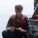 A young male sailor or naval personnel is crouched on the deck of a military vessel at sea, working with electrical cables or connectors. He is wearing a red life vest over a dark blue uniform, consistent with naval working attire. The ocean is visible in the background under an overcast sky.
