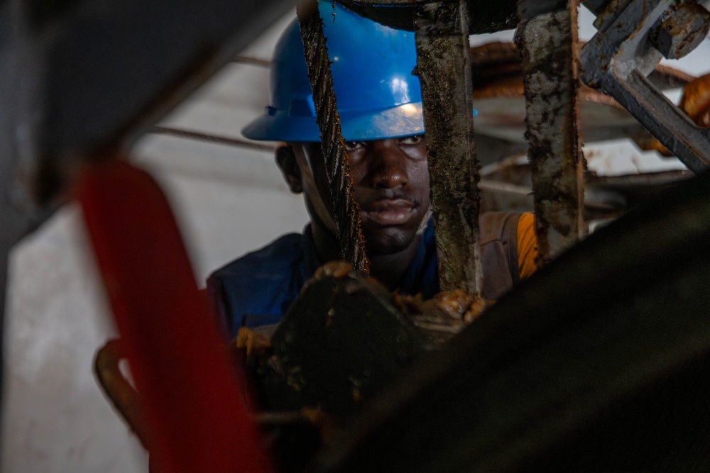 A male worker wearing a blue hard hat and blue work shirt is photographed in close proximity through a gap in heavy industrial equipment. His face and hands are visibly soiled with dark grease or mud, suggesting active manual labor in a demanding environment. The surrounding machinery appears heavily corroded and worn, indicating an industrial or construction setting.