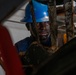 A male worker wearing a blue hard hat and blue work shirt is photographed in close proximity through a gap in heavy industrial equipment. His face and hands are visibly soiled with dark grease or mud, suggesting active manual labor in a demanding environment. The surrounding machinery appears heavily corroded and worn, indicating an industrial or construction setting.