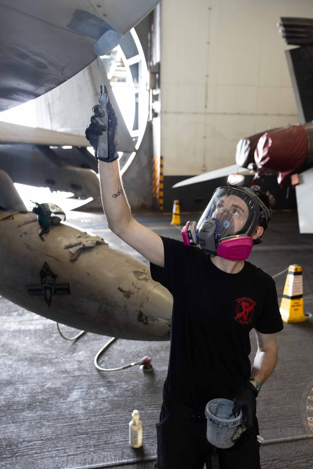 A technician wearing a full-face respirator mask and black gloves is painting or applying a coating to the underside of a military aircraft in what appears to be a hangar bay. The worker holds a brush in one raised hand and a paint bucket in the other, working on the aircraft's surface. The setting appears to be a naval or military aviation maintenance facility.