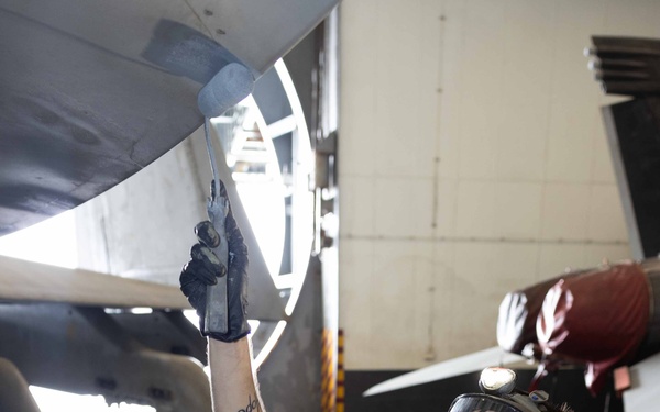 A technician wearing a full-face respirator mask and black gloves is painting or applying a coating to the underside of a military aircraft in what appears to be a hangar bay. The worker holds a brush in one raised hand and a paint bucket in the other, working on the aircraft's surface. The setting appears to be a naval or military aviation maintenance facility.