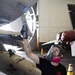 A technician wearing a full-face respirator mask and black gloves is painting or applying a coating to the underside of a military aircraft in what appears to be a hangar bay. The worker holds a brush in one raised hand and a paint bucket in the other, working on the aircraft's surface. The setting appears to be a naval or military aviation maintenance facility.