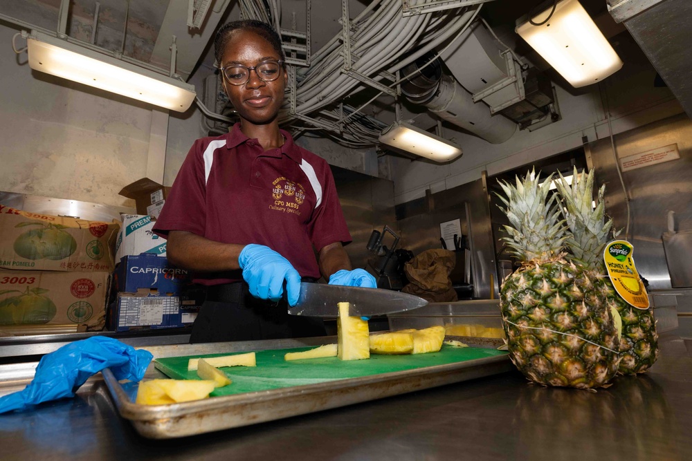 A person wearing a maroon polo shirt with 'CPO MESS Culinary Specialist' insignia and blue latex gloves is cutting pineapple in what appears to be a military ship's galley kitchen. Pineapple slices are arranged on a green cutting board atop a metal tray, with a whole pineapple labeled 'Honey Pineapple' by Sunpride visible to the right. The industrial ceiling with exposed pipes and conduits is consistent with a naval vessel interior.