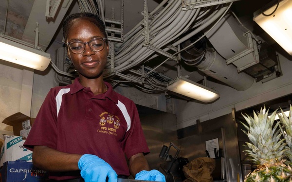 A person wearing a maroon polo shirt with 'CPO MESS Culinary Specialist' insignia and blue latex gloves is cutting pineapple in what appears to be a military ship's galley kitchen. Pineapple slices are arranged on a green cutting board atop a metal tray, with a whole pineapple labeled 'Honey Pineapple' by Sunpride visible to the right. The industrial ceiling with exposed pipes and conduits is consistent with a naval vessel interior.