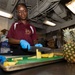 A person wearing a maroon polo shirt with 'CPO MESS Culinary Specialist' insignia and blue latex gloves is cutting pineapple in what appears to be a military ship's galley kitchen. Pineapple slices are arranged on a green cutting board atop a metal tray, with a whole pineapple labeled 'Honey Pineapple' by Sunpride visible to the right. The industrial ceiling with exposed pipes and conduits is consistent with a naval vessel interior.