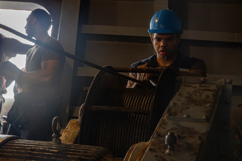Two workers are operating heavy industrial machinery, likely a cable winch or hoisting mechanism in what appears to be a ship or industrial facility. The foreground worker is wearing a blue hard hat and has a whistle in their mouth, suggesting a supervisory or signaling role during the operation. The environment is dimly lit with a bright light source visible in the background.