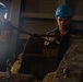 Two workers are operating heavy industrial machinery, likely a cable winch or hoisting mechanism in what appears to be a ship or industrial facility. The foreground worker is wearing a blue hard hat and has a whistle in their mouth, suggesting a supervisory or signaling role during the operation. The environment is dimly lit with a bright light source visible in the background.