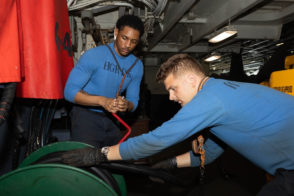 Two individuals wearing matching blue sweatshirts are working together on equipment in what appears to be a naval vessel's interior workspace. One person is leaning over and handling a large green cylindrical object while wearing black gloves, while the other observes and assists. The environment shows industrial naval infrastructure including pipes, cables, and overhead lighting.