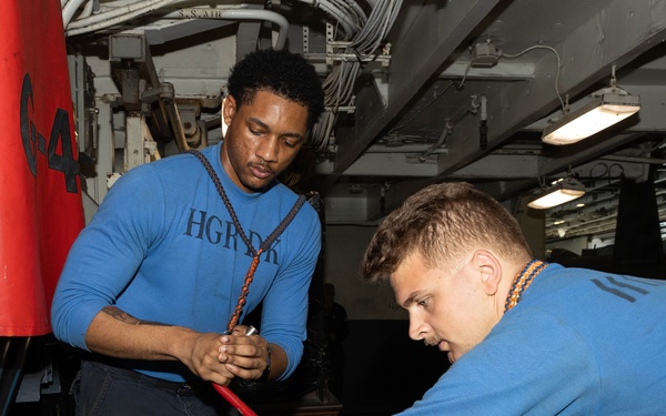 Two individuals wearing matching blue sweatshirts are working together on equipment in what appears to be a naval vessel's interior workspace. One person is leaning over and handling a large green cylindrical object while wearing black gloves, while the other observes and assists. The environment shows industrial naval infrastructure including pipes, cables, and overhead lighting.