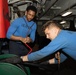 Two individuals wearing matching blue sweatshirts are working together on equipment in what appears to be a naval vessel's interior workspace. One person is leaning over and handling a large green cylindrical object while wearing black gloves, while the other observes and assists. The environment shows industrial naval infrastructure including pipes, cables, and overhead lighting.