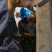 A worker wearing a blue hard hat and safety vest is visible in a confined or industrial space, partially obscured by structural elements in the foreground. The worker appears to be handling tools or equipment with gloved hands. The setting suggests an industrial, construction, or maintenance environment.