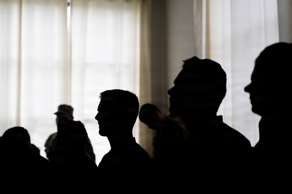 The image shows a group of people rendered as dark silhouettes against a bright, backlit window with sheer curtains. The strong contrast between the dark foreground figures and the bright background creates a dramatic, high-contrast composition. Individual facial features are indistinguishable, though head shapes and profiles are visible.