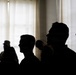 The image shows a group of people rendered as dark silhouettes against a bright, backlit window with sheer curtains. The strong contrast between the dark foreground figures and the bright background creates a dramatic, high-contrast composition. Individual facial features are indistinguishable, though head shapes and profiles are visible.
