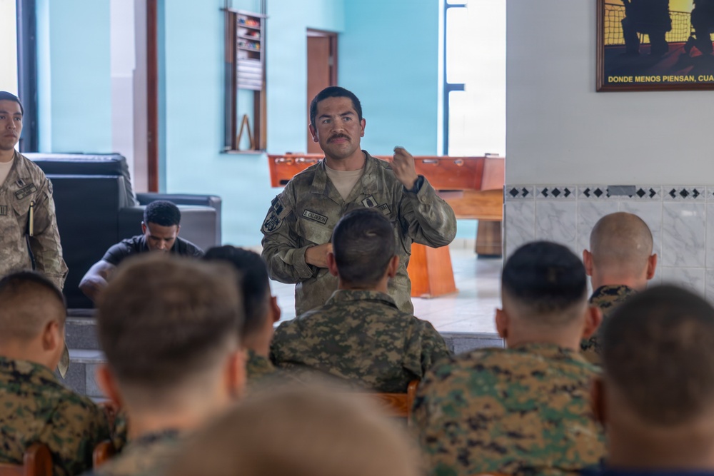A military briefing or instruction session is taking place indoors, with one soldier in US Army camouflage uniform standing and addressing a seated audience of multiple soldiers wearing woodland digital camouflage uniforms. The standing speaker appears to be a non-commissioned or commissioned officer, identifiable by rank insignia on his uniform. The setting appears to be a recreational or common area of a military facility, with a foosball table visible in the background.