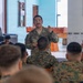 A military briefing or instruction session is taking place indoors, with one soldier in US Army camouflage uniform standing and addressing a seated audience of multiple soldiers wearing woodland digital camouflage uniforms. The standing speaker appears to be a non-commissioned or commissioned officer, identifiable by rank insignia on his uniform. The setting appears to be a recreational or common area of a military facility, with a foosball table visible in the background.
