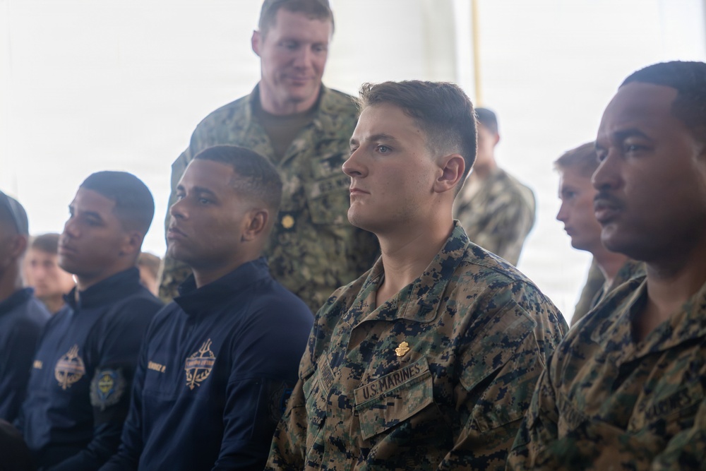 A group of U.S. military personnel are seated and standing in what appears to be a briefing or assembly setting under a tent or canopy. The individuals are wearing a mix of U.S. Marine Corps MARPAT camouflage uniforms and Navy blue athletic/service uniforms. The central figure is clearly identifiable as a U.S. Marine based on the visible 'U.S. MARINES' tape on his uniform.