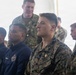 A group of U.S. military personnel are seated and standing in what appears to be a briefing or assembly setting under a tent or canopy. The individuals are wearing a mix of U.S. Marine Corps MARPAT camouflage uniforms and Navy blue athletic/service uniforms. The central figure is clearly identifiable as a U.S. Marine based on the visible 'U.S. MARINES' tape on his uniform.