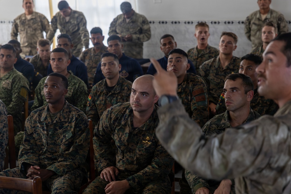 A military briefing or instruction session is taking place indoors, with a large group of soldiers seated and attentively listening to an instructor or speaker on the right side of the frame. The attendees appear to wear a mix of camouflage uniforms, including what appears to be USMC digital camouflage and other patterns, suggesting a multinational or joint-force training exercise. The speaker is gesturing with one hand, indicating an active instructional or Q&amp;A moment.