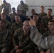 A military briefing or instruction session is taking place indoors, with a large group of soldiers seated and attentively listening to an instructor or speaker on the right side of the frame. The attendees appear to wear a mix of camouflage uniforms, including what appears to be USMC digital camouflage and other patterns, suggesting a multinational or joint-force training exercise. The speaker is gesturing with one hand, indicating an active instructional or Q&amp;A moment.