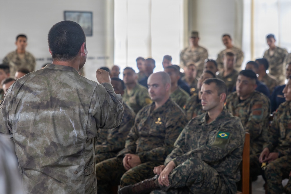 A military briefing or instruction session is taking place indoors, with one uniformed individual addressing a seated group of soldiers. At least one soldier in the audience is visibly wearing a Brazilian flag patch on their uniform sleeve, indicating a multinational military gathering. The speaker stands with their back to the camera, gesturing with a raised fist toward the attentive audience.