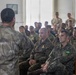 A military briefing or instruction session is taking place indoors, with one uniformed individual addressing a seated group of soldiers. At least one soldier in the audience is visibly wearing a Brazilian flag patch on their uniform sleeve, indicating a multinational military gathering. The speaker stands with their back to the camera, gesturing with a raised fist toward the attentive audience.