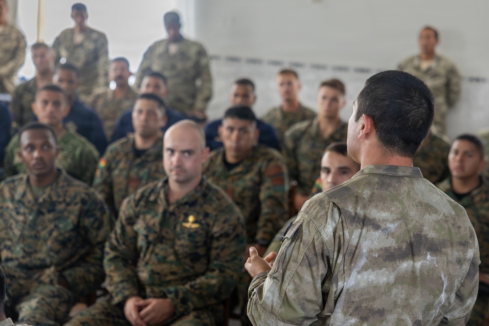 A military briefing or instruction session is taking place indoors, with one uniformed individual addressing a large group of seated soldiers. The audience consists of personnel wearing multiple different camouflage patterns, suggesting a multinational or joint military exercise. The speaker is photographed from behind, facing the assembled group in what appears to be a classroom or meeting room setting.