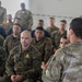 A military briefing or instruction session is taking place indoors, with one uniformed individual addressing a large group of seated soldiers. The audience consists of personnel wearing multiple different camouflage patterns, suggesting a multinational or joint military exercise. The speaker is photographed from behind, facing the assembled group in what appears to be a classroom or meeting room setting.