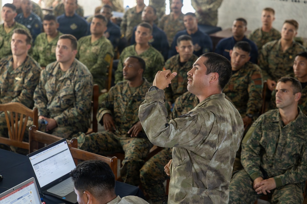 A military briefing or training session is taking place in an indoor venue with a large group of soldiers seated as an audience. A soldier in a distinctive camouflage pattern (different from the majority) appears to be speaking or asking a question with a raised hand gesture. Multiple camouflage uniform patterns are visible, suggesting a multinational or joint military exercise.