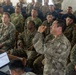 A military briefing or training session is taking place in an indoor venue with a large group of soldiers seated as an audience. A soldier in a distinctive camouflage pattern (different from the majority) appears to be speaking or asking a question with a raised hand gesture. Multiple camouflage uniform patterns are visible, suggesting a multinational or joint military exercise.