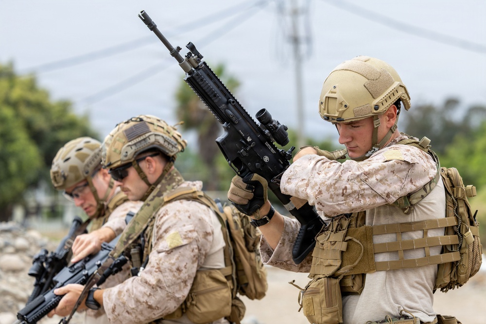 Three military personnel in tactical gear are shown in what appears to be a training or operational environment. The foreground soldier is inspecting or handling an assault rifle, while the others behind him are similarly equipped. The personnel are wearing a mix of MARPAT (Marine pattern) and MultiCam camouflage uniforms with full tactical kit.