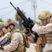 Three military personnel in tactical gear are shown in what appears to be a training or operational environment. The foreground soldier is inspecting or handling an assault rifle, while the others behind him are similarly equipped. The personnel are wearing a mix of MARPAT (Marine pattern) and MultiCam camouflage uniforms with full tactical kit.