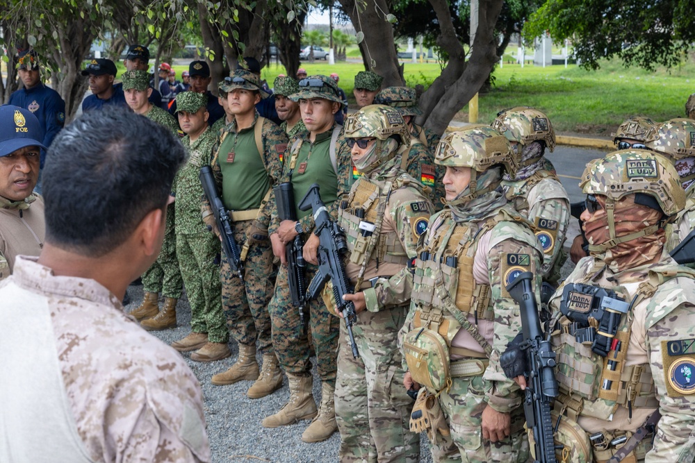 A multinational military gathering showing personnel in various uniforms and equipment levels, ranging from lightly equipped troops in camouflage fatigues to heavily geared operators in full tactical kit with helmets and body armor. Several soldiers are carrying assault rifles and appear to be receiving a briefing from an individual in desert camouflage facing away from the camera. Shoulder patches visible on some personnel suggest Bolivian military affiliation among others.