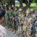 A multinational military gathering showing personnel in various uniforms and equipment levels, ranging from lightly equipped troops in camouflage fatigues to heavily geared operators in full tactical kit with helmets and body armor. Several soldiers are carrying assault rifles and appear to be receiving a briefing from an individual in desert camouflage facing away from the camera. Shoulder patches visible on some personnel suggest Bolivian military affiliation among others.
