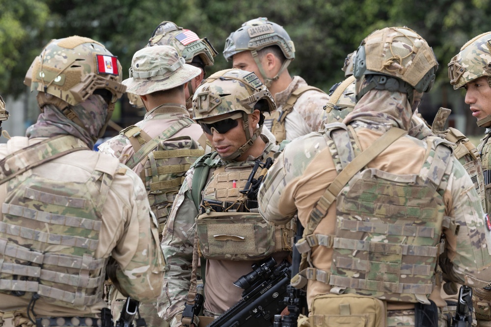 A group of military personnel in full tactical gear are gathered in what appears to be a briefing or coordination huddle. Multiple nationalities are represented, as evidenced by visible flag patches including a US flag and what appears to be a Peruvian flag. The soldiers are equipped with helmets, body armor, tactical vests, and weapons.