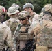 A group of military personnel in full tactical gear are gathered in what appears to be a briefing or coordination huddle. Multiple nationalities are represented, as evidenced by visible flag patches including a US flag and what appears to be a Peruvian flag. The soldiers are equipped with helmets, body armor, tactical vests, and weapons.