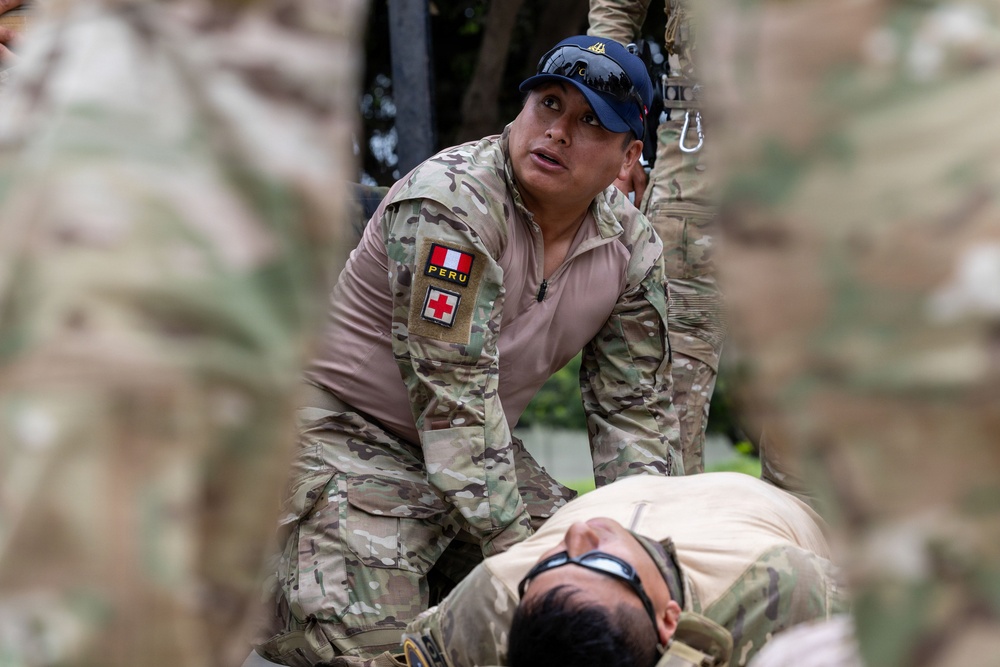 A Peruvian military medic in camouflage uniform is attending to a person lying on the ground, appearing to perform or simulate a medical/casualty care procedure. The soldier wears a patch on the left sleeve identifying 'PERU' with a Red Cross symbol, indicating medical personnel. The scene appears to be a training exercise or field medical response scenario.