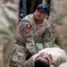 A Peruvian military medic in camouflage uniform is attending to a person lying on the ground, appearing to perform or simulate a medical/casualty care procedure. The soldier wears a patch on the left sleeve identifying 'PERU' with a Red Cross symbol, indicating medical personnel. The scene appears to be a training exercise or field medical response scenario.