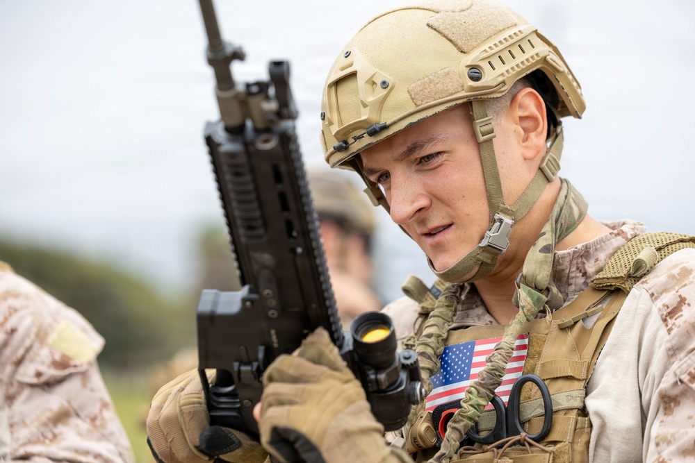 A young male soldier in full tactical gear is holding a rifle and appears to be in a field environment. He is wearing a tan ballistic helmet, multicam/OCP patterned uniform, and a tactical vest with a visible American flag patch on the chest. The image is a close-up portrait-style photograph with a blurred background.