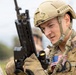 A young male soldier in full tactical gear is holding a rifle and appears to be in a field environment. He is wearing a tan ballistic helmet, multicam/OCP patterned uniform, and a tactical vest with a visible American flag patch on the chest. The image is a close-up portrait-style photograph with a blurred background.