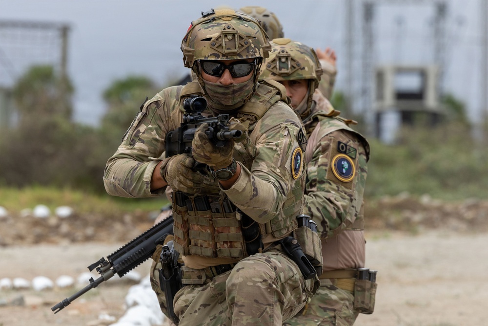 Two heavily equipped military or special operations personnel are shown in a tactical formation, with the foreground soldier aiming a rifle directly toward the camera. Both individuals wear multicam camouflage uniforms, tactical plate carriers, and ballistic helmets with accessory mounts. The setting appears to be an outdoor training or operational environment with an industrial structure visible in the background.
